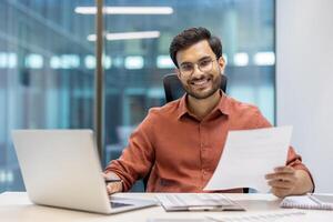 Confident man in office setting working with laptop and document. Smiling while handling tasks. Demonstrates professionalism, focus, and positive attitude at work. photo
