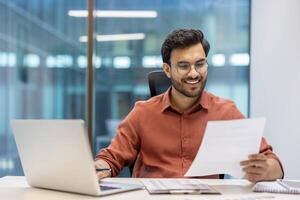 Confident man in office reading document while seated at desk with laptop and notebook, showcasing a positive and productive work atmosphere. Professional setting highlights engagement in business photo