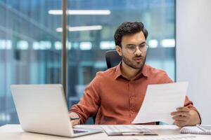 Businessman in office analyzing documents and working on laptop. Focused professional handling tasks with efficiency and concentration. Corporate environment facilitating dedicated workday activities photo