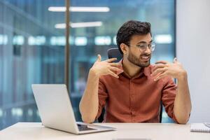 Business professional feeling discomfort due to heat in office. Young man gesturing frustration while working at desk with laptop. Overheated workplace causing stress and communication challenges. photo