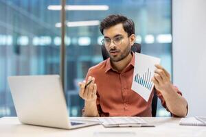 Businessman in office holding paper chart, explaining financial data during call on laptop. Focus on analysis, communication, technology. Reflects professional environment and office work photo