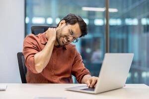 Office employee feeling neck pain while working on laptop. Professional in modern office showing signs of stress and discomfort, highlighting ergonomic issues common in workplace environments. photo