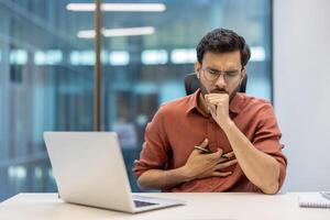 Businessman in office feeling unwell, hand on chest while coughing. Depicts stress, health issue in professional setting. Workplace with laptop, suggests need for attention to employee well-being. photo