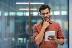 Serious programmer focused on tablet screen in modern office. Individual wearing glasses and casual attire analyzing information to solve problems or develop software. photo