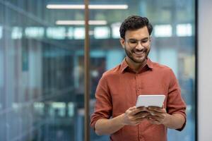 Smiling programmer tests application on tablet in modern office. Casual professional wears glasses and brown shirt, demonstrating focus and tech expertise. photo
