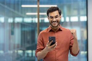Happy businessman in office celebrating success while viewing smartphone screen. Shows delight and achievement in workplace setting. Captures modern business atmosphere and motivation person photo