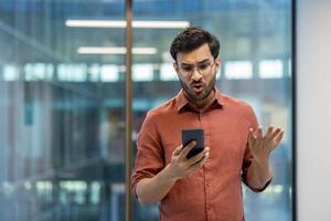 Man in office holding smartphone with confused expression, gesturing frustration. Business professional dealing with unexpected news or technical issues, showcasing emotions of surprise and disbelief photo