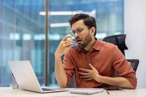 Man in office experiencing asthma symptoms using inhaler for relief. feeling discomfort. Scene portrays health issues in workplace setting, focusing on respiratory care and stress management. photo