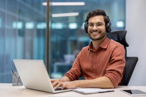 Young professional with glasses and headphones using laptop in modern office setting. Person smiling confidently while engaged in digital work. Office environment encourages creativity and focus photo