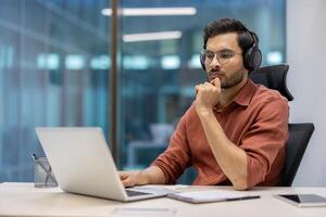 Serious businessman in modern office setting wearing headphones working on laptop. Focused on tasks, demonstrates concentration and professionalism. Represents technology use in business photo