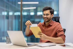 Young professional using laptop, opening envelope with joyful expression. Workplace setting with personal items, casual attire. Concept of work satisfaction, communication, modern technology. photo
