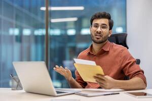 Office worker looks distressed after opening unexpected mail. Seated at desk with laptop and phone, expression reflects surprise and concern. Scene captures challenging moment in professional setting photo
