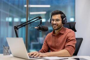 Smiling man wearing headphones recording audio podcast with professional microphone setup and laptop in modern office. Engaged in work, representing creativity, technology, and media production industry. photo