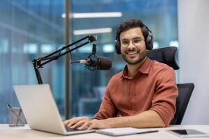 Young man smiling while recording podcast in modern office. Wearing headphones, speaking into microphone. Laptop open in front, capturing engaged expression. photo