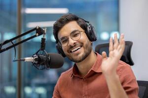 Smiling man in studio speaks into microphone wearing headphones, creating podcast. Friendly wave towards camera suggests engaging interaction. photo