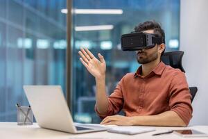 Young professional in modern office using virtual reality headset while interacting with laptop for innovative experience. Person explores new technology concepts, enhancing digital interaction photo