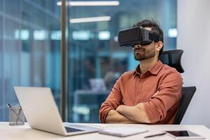 Man in office wearing virtual reality headset, exploring immersive technology. Seated at desk with laptop. Concept includes technology, innovation, and a futuristic work environment. photo