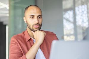Young man thinking while sitting at desk inside office. Businessman close up working with laptop, looking at screen, reading electronic report. photo