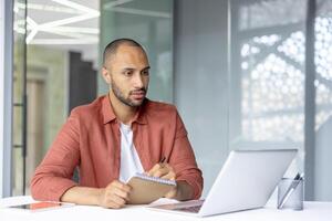 Serious thinking man writing data in notebook, employee watching training webinar, online business meeting, remote conference. Businessman working with laptop for call inside office. photo