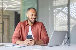 Successful joyful man with phone in hands smiling working inside office with laptop. Company employee satisfied with results of achievement at workplace. photo