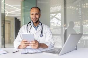 Successful experienced doctor sitting at desk inside the clinic office and using a tablet computer. Man in a white medical coat, using an application for online communication and consulting patients. photo