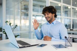 A cheerful young man with curly hair waves while engaging in a call in a bright, modern office environment. The frame captures a sense of friendliness and connectivity in the corporate world. photo