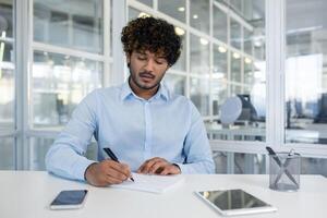 Young professional male in a modern office setting, intently writing in a notebook with a smartphone and computer nearby. Represents productivity and workplace concentration. photo