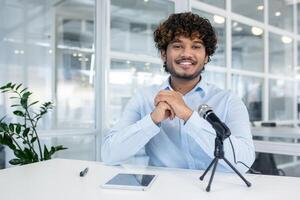 Confident businessman sitting at an office desk with a microphone, ready for a podcast interview. Office setting with modern decor in the background. photo
