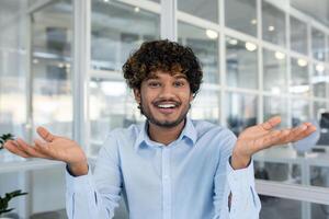 Cheerful young adult in a light blue shirt gesturing with hands, expressing positivity and confidence in a bright office setting. photo