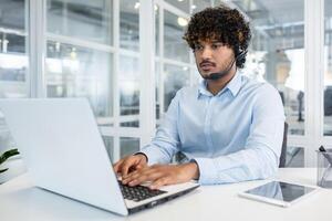 A young, professional man wearing a headset focuses intently on his laptop in a modern office environment, suggesting productivity and business communication. photo