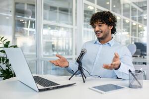 An enthusiastic young man in a modern office space is engaging with an online audience via microphone and laptop. He displays a lively expression and gestures. photo