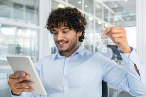 A cheerful young man with curly hair, showcasing a house keychain while browsing on his tablet in a bright office setting. photo