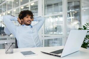 A young businessman relaxes at his desk with hands behind his head, looking thoughtful. The modern office setting with bright lighting encapsulates a moment of calm and contemplation during a busy workday. photo