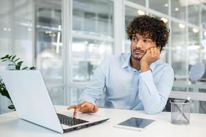 A young professional appears bored and disengaged while working at his laptop in a bright, modern office. He gazes out a window, seemingly lost in thought. photo