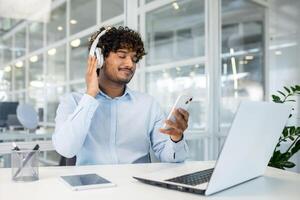 A young man with curly hair is listening to music on his headphones while holding a smartphone in a bright office environment. He appears relaxed and content with his work setup. photo
