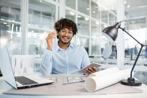 A cheerful male architect with curly hair working in a contemporary office. He holds a pen tablet, smiles brightly, and gestures with satisfaction towards his architectural blueprints. photo