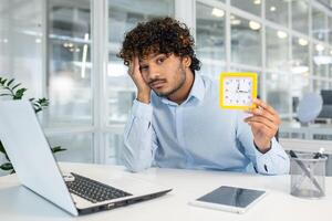 A young man sits at his office desk, displaying signs of boredom and lack of motivation as he holds a clock, signaling the slow passage of time. photo