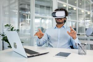 Curly-haired young professional immersed in virtual reality using a VR headset while engaging with invisible interfaces at his workplace equipped with various modern devices. photo