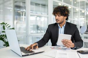 A focused young man multitasks with a laptop and tablet at a desk surrounded by light-filled modern office space. He exudes professionalism and efficiency in a corporate environment. photo