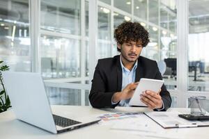 A focused young businessman analyzes data on a tablet while sitting at a desk in a bright, contemporary office. Essential tools and documents surround him. photo