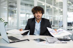 Frustrated indian male in jacket over shirt sitting by loaded table and spreading hands with documents. Young manager feeling stressed with paperwork and looking at camera with angry expression. photo