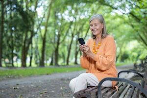Senior woman seated on park bench using smartphone, smiling and enjoying outdoor leisure. Wearing casual , showing happiness and connectivity in nature setting with lush greenery. photo