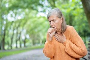 Elderly woman coughing in outdoor park setting showing discomfort. Holding chest with serious expression. Scene captures health issues, age, and lifestyle concerns. Green leafy trees in background. photo