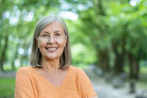Mature woman smiles confidently standing outside . Image captures essence of positive aging and happiness. Trees and greenery create serene backdrop for this lifestyle portrait of an older adult. photo
