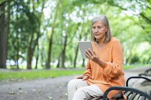 Older woman seated on park bench using tablet, engaging with digital technology. Outdoor setting emphasizes tranquil lifestyle, blending modern technology with nature. photo