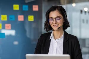 Businesswoman wearing headset smiling while working on laptop computer. Customer service agent or support representative in office. Communication and technology concepts woman using phone. photo