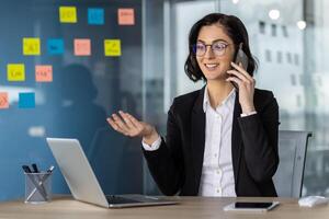 Confident businesswoman in glasses having phone call in modern office setting. Analyzing charts on laptop, smiling while discussing business strategies. Productivity and professionalism concept photo