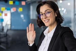 Confident businesswoman waving during call in modern office. She wears stylish glasses and blazer, showcasing professionalism and communication skills while holding phone. photo