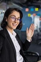 Confident businesswoman wearing glasses waves in a modern office setting. Represents communication, networking, and professionalism in a business environment. Suitable for business themes. photo