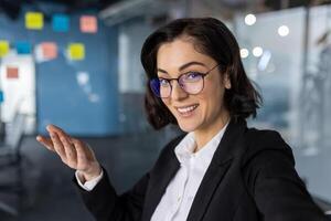 Businesswoman boss using phone for call, smiling at camera, gesturing confidently. Professional office setting with colorful notes background. Ideal for concepts of communication, technology. photo